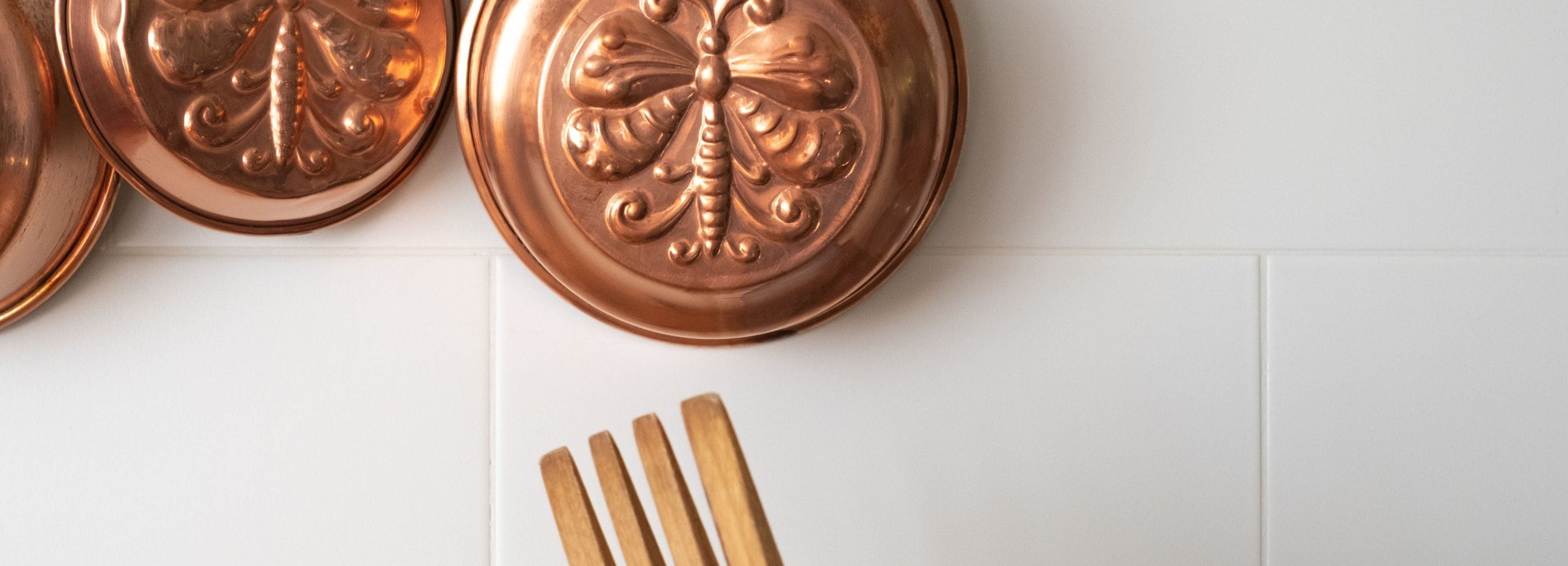 Copper pots hanging on a rack with wooden and black utensils in a holder against a white tiled wall.